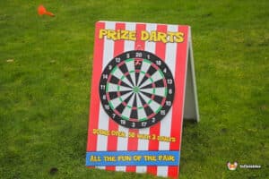 A colorful prize darts game board with a dartboard design stands on green grass, advertising the rules for scoring and fun.