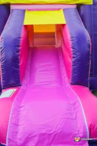 A vibrant purple and pink inflatable bouncer featuring a slide, set up for children's play in an outdoor area.