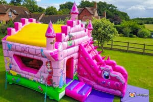 A pink bouncy castle featuring a slide, with a princess hand painted artwork, set in a fairytale theme with a dragon slide.