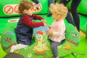 Two young girls happily seated on a green double rocker designed as an animal, engaged in playful interaction.