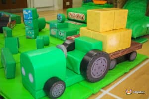 A play area showcasing a green tractor with a trailer, set against a backdrop of grass and colorful play structures.