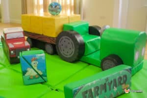 A vibrant play area with a green tractor pulling a trailer, surrounded by grass and various play equipment.