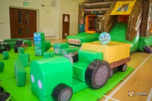 A play area containing a green tractor with a trailer, situated on grass with nearby play equipment for children.