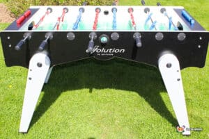 A foosball table with a black and white design, featuring red and blue players, sits on a grassy lawn under bright sunlight.