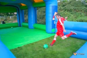 A man in red kicks a Football ball inside a colourful inflatable bouncy castle testing his speed.