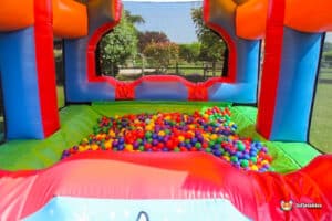 A colourful inflatable gigantic ball pool filled with a variety of plastic balls, set in a sunny outdoor area with greenery in the background.