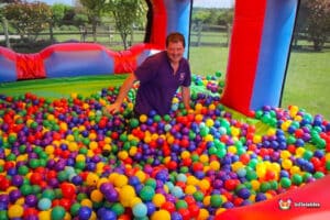 A person sits in a colorful ball pit inside a bouncy castle, surrounded by vibrant red, blue, yellow, green, and purple balls.
