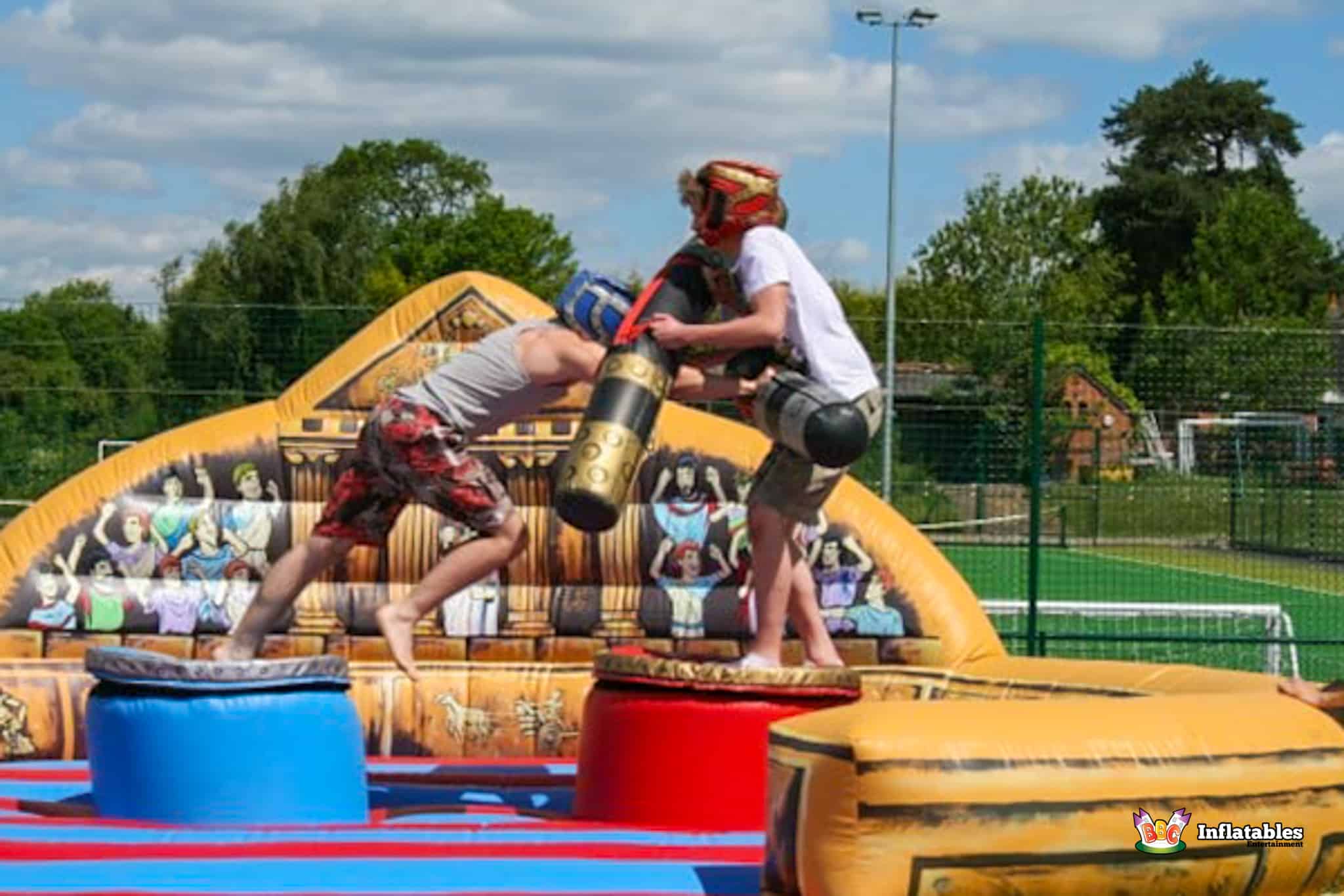 Two boys participating in a gladiator duel on an inflatable, energetically swinging foam sticks at each other.