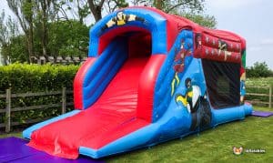 A colourful inflatable hero fun run featuring superheroes, with a large red slide and mesh panels, set in a grassy outdoor area.