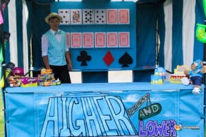 A side stall featuring a colourful banner reading "HIGHER AND LOWER," with playing cards displayed on a backdrop and prizes on the table.