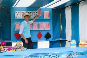 A magician in a blue and white striped vest performs card tricks at a colorful carnival booth filled with toys and party supplies.