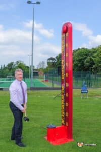 A man stands beside a large red high striker, resembling a thermometer, at a carnival game.