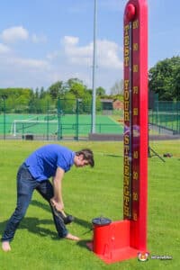 A man stands next to a tall red high striker, designed to resemble a thermometer, at a festive event.