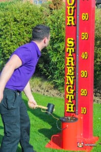 A man poses next to a large red high striker, which looks like a thermometer, at a fairground.