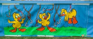 Three playful cartoon ducks hang from colorful hooks against a bright blue backdrop, evoking a cheerful, whimsical atmosphere.