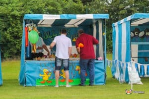 Two men play a carnival game Hook a duck side stall at a colourful striped booth featuring rubber ducks and prizes, set in a grassy outdoor area.
