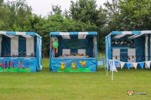 Three colorful fair stalls labeled "Hupla," "Roll Up! Roll Up!," and "Coconut" are set up in a grassy area, surrounded by trees.