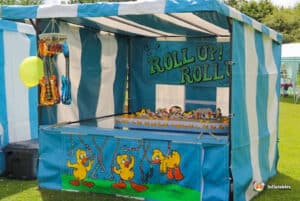 A colourful carnival hook a duck side stall featuring "Roll Up! Roll!" signage, playful duck graphics, and inflatable guitars, set in a festive outdoor space.