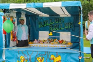 A colorful carnival booth with a sign saying "Roll Up! Roll Up!", featuring games and prizes, including rubber ducks and stuffed toys.