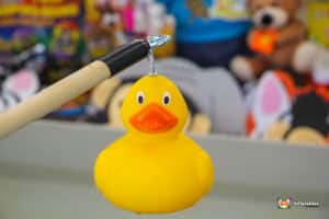 A bright yellow rubber duck hanging from a fishing pole, with plush toys blurred in the background.