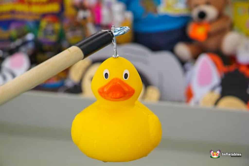 A bright yellow rubber duck hanging from a fishing pole, with plush toys blurred in the background.