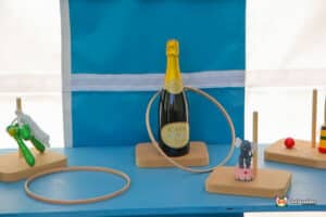 A bottle of Cava champagne is displayed with wooden toys and rings on a blue table, set against a blue and white backdrop.