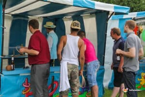 A group of people stand in front of a colorful stall with games and drinks, engaging in a fun outdoor event.