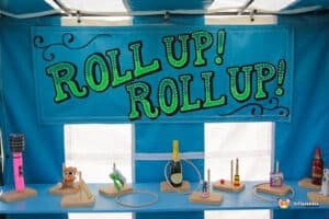 A vibrant banner reads "Roll Up! Roll Up!" over a booth displaying various playful competition prizes like toys and bottles.