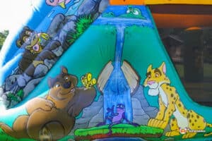 A colorful inflatable slide featuring cartoon animals: a bear, frog, and leopard, with a waterfall backdrop in a vibrant outdoor setting.