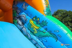 A colorful inflatable slide features a cartoon alligator waving, surrounded by water and greenery against a bright blue sky.