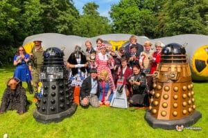 A group dressed in various costumes stands beside two iconic Dalek figures in a vibrant outdoor setting.