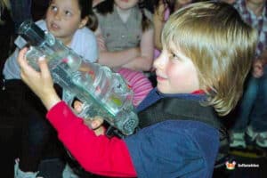 A child holds a clear toy laser gun, focused intently on their surroundings, with excited peers visible in the background.