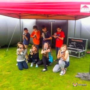A group of children posing playfully under a red tent, holding toy guns in a grassy field, with inflatable structures in the background.