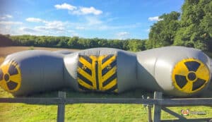 A large inflatable Laser Tag Arena shaped like a nuclear barrel, decorated with radioactive symbols, set in a grassy field under a blue sky.