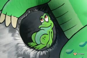 Cartoon frog peeks out from a hole in a colorful, large inflatable, surrounded by bright summer scenery.