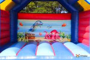 A colourful inflatable bouncy castle featuring a dolphin, octopus, and fish against a beach backdrop, with bright red and blue walls.