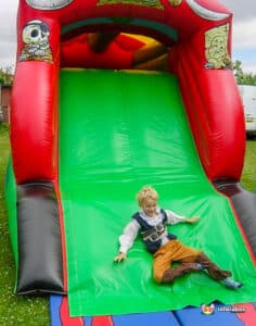 A colorful inflatable slide in a grassy area, with a child dressed in a pirate costume sitting on the green sliding surface.