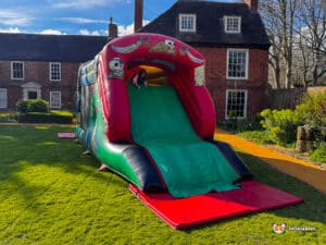 A child climbs a colorful inflatable slide in a grassy yard, with a red-brick house and trees in the background under a blue sky.
