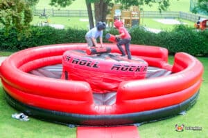 Two boys playfully spar on an inflatable gladiator duel, surrounded by bright colors and excitement.