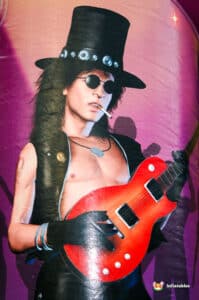 An electric guitarist poses confidently, wearing a tall black hat and a sleeveless leather vest, holding a vibrant red guitar.