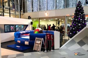 A festive indoor setting features a blue inflatable bull ride with a scoring board, surrounded by holiday decorations and a Christmas tree.