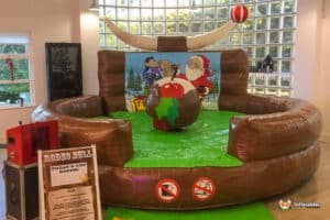 A festive rodeo bull attraction with a holiday theme, featuring Santa and cartoon characters, surrounded by a safe inflatable barrier.