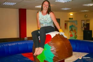 A person wearing gloves sits atop a large, inflatable chocolate cake in a festive indoor setting. Colorful decorations in the background.
