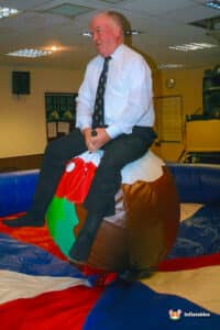 A person in a white shirt and tie sits atop a large, inflatable Christmas pudding in a festive indoor setting, surrounded by colorful mats.