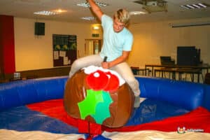 A young person rides a mechanical bull designed to look like a festive Christmas pudding, surrounded by a safety inflatable ring.