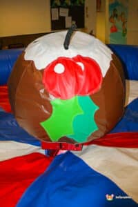 A large inflatable Christmas pudding with red berries and green leaves sits on colorful matting in a festive indoor setting.