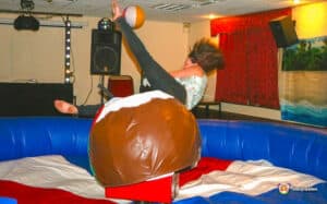 A young woman tumbles off a mechanical bull during a playful moment at a party, surrounded by colorful inflatable barriers.