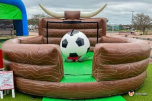 Inflatable bull riding game designed for football, featuring a large soccer ball and bullhorn decor, set on green turf.