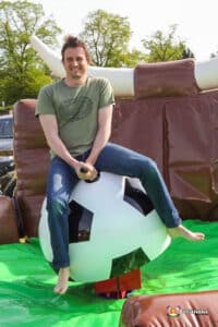 A person enjoys a ride on a mechanical football ball surrounded by inflatable barriers, set outdoors on a sunny day.