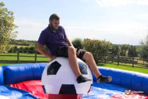 A person sits on a large inflatable football ball in a colorful bouncy arena, surrounded by greenery and a blue sky.